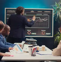 A presenter writes on a large digital screen displaying a dark, complex annual statistics graph during a business meeting