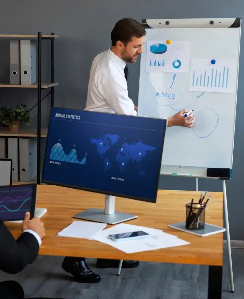 A businessman presents to a colleague, drawing on a whiteboard next to a monitor displaying a world map and line graphs