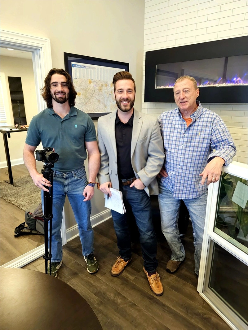 Three men stand together in a modern office setting, featuring a stylish fireplace and a wall map in the background.