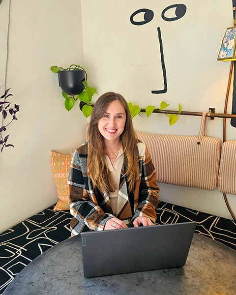 A woman in a patterned jacket types on a laptop at a cozy, decorated workspace with greenery and abstract wall art.